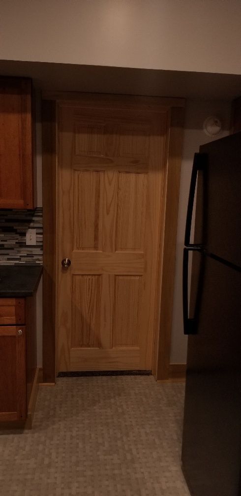 Wooden door in a kitchen setting with brown cabinets and a black refrigerator.