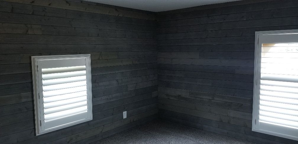 A room with gray wood panel walls and two windows with white shutters.
