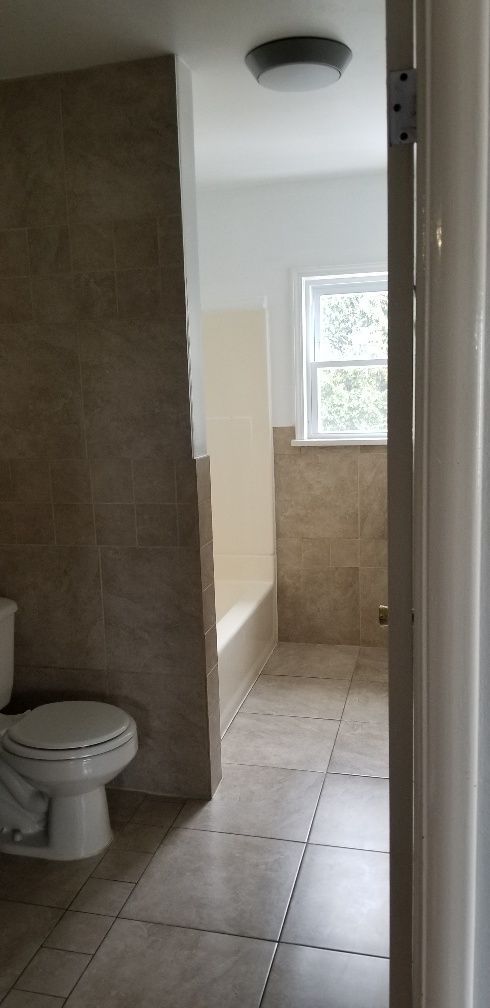 Bathroom interior with toilet, bathtub, and window. Beige tile on the floor and walls. A flush-mount ceiling light is visible.