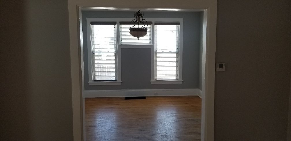 Interior view: Room with wood floors, two windows with blinds, and a chandelier, seen through a doorway.