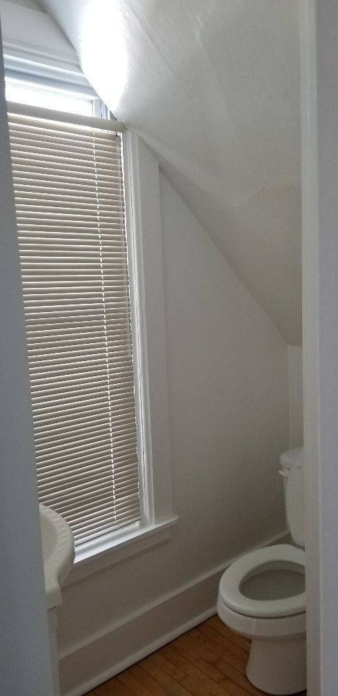 Small bathroom with toilet and window with blinds. Angled ceiling and wood flooring.