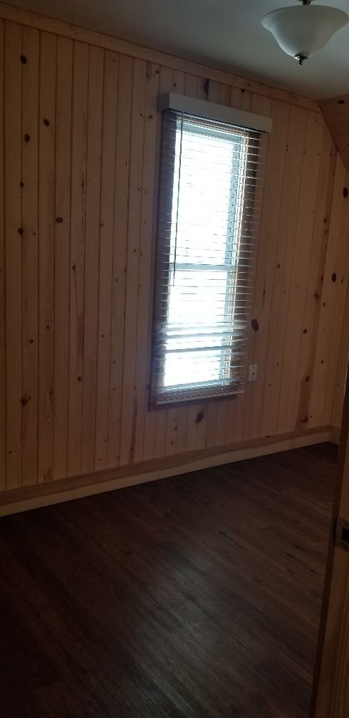 Empty room with wooden walls and floor, a window with blinds, and a ceiling light.