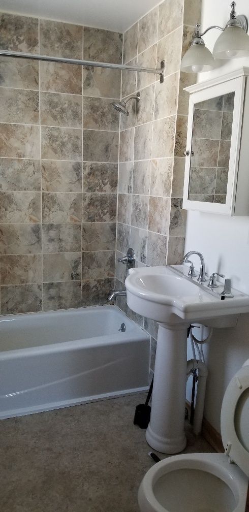 A bathroom with a white tub, pedestal sink, toilet, and tiled walls. A shower curtain rod is above the tub.
