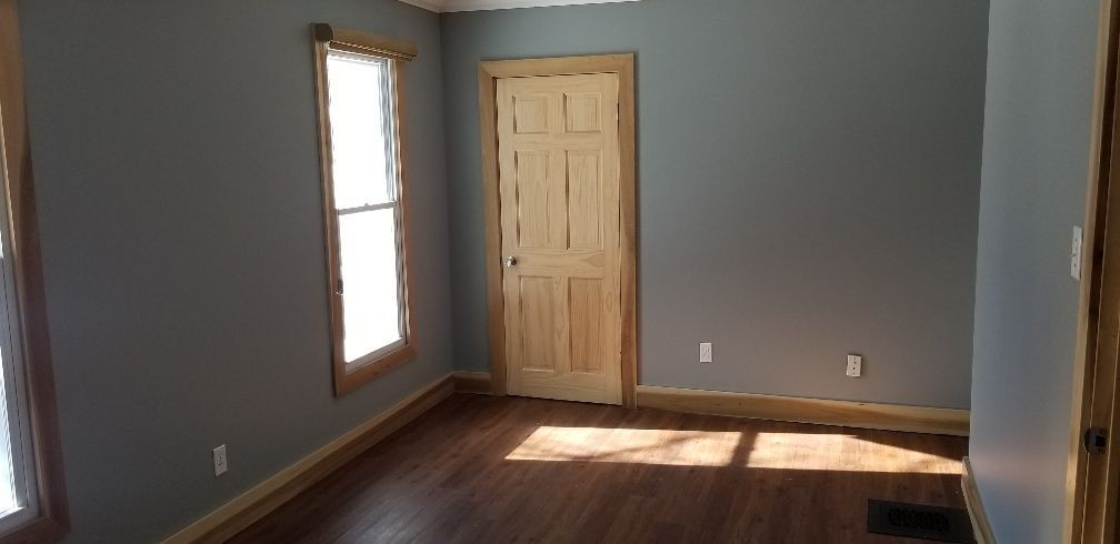 An empty room with gray walls, wood trim, and a closed door. Sunlight streams through a window.