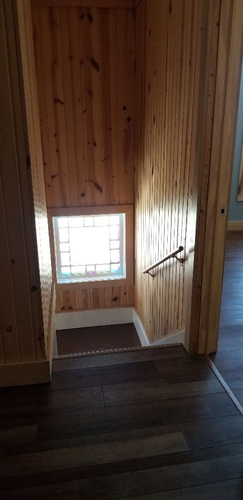 Staircase with wooden paneling and a small window. Handrail on the right, dark flooring in the foreground.
