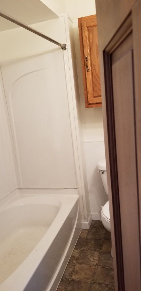 Bathroom with a white bathtub, toilet, and wooden cabinet. Brown floor, white walls.