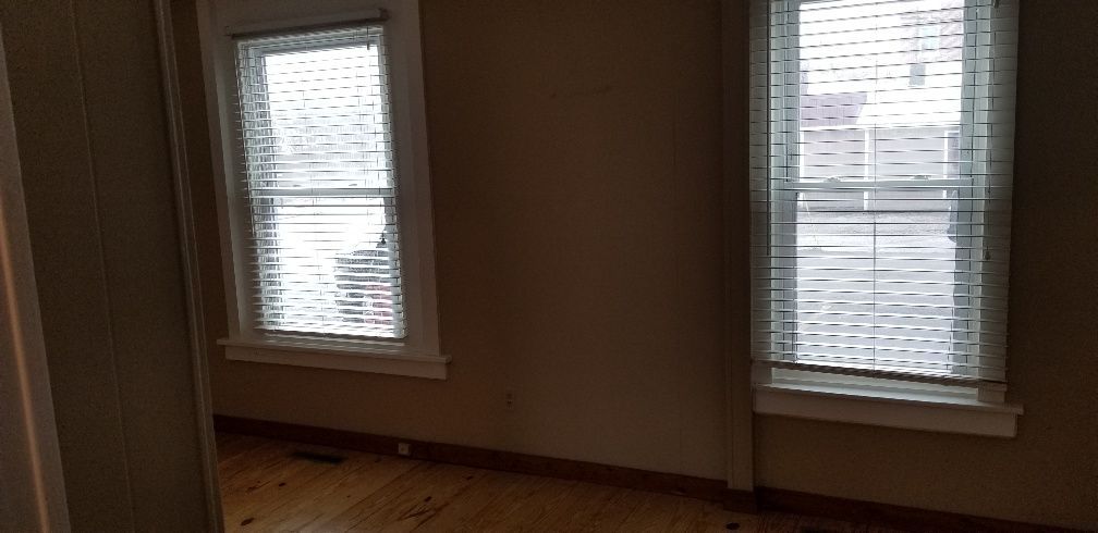 Empty room with two windows, light blinds, beige walls, and wood floor.