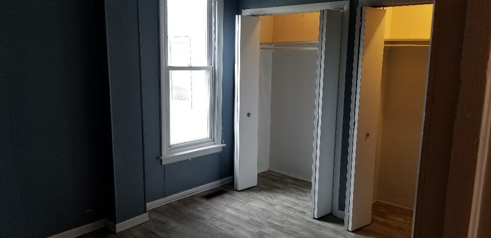 A small bedroom with two closets and a window. The walls are blue and the floor has a wood grain look.