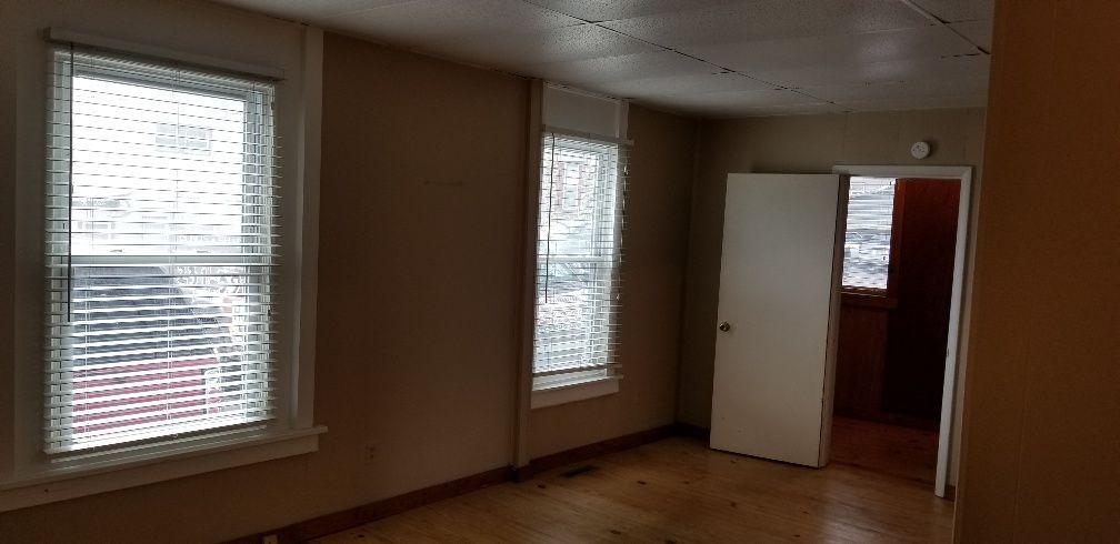Empty room with two windows and a doorway. Beige walls, wooden floor, blinds on windows.