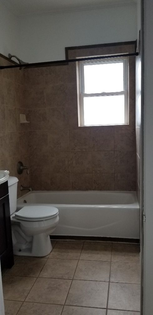 Bathroom with a toilet, bathtub, and window. Beige tile flooring and wall tile.