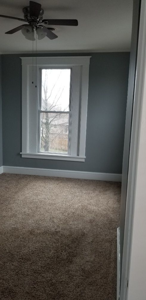 Empty bedroom with gray walls, white trim, and brown carpet. Window visible.