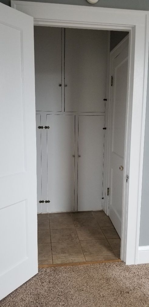 Open white door revealing a white closet with multiple cabinets; a light tan tiled floor meets the beige carpet.