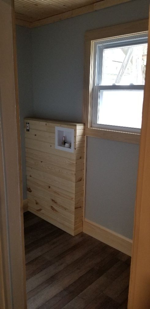Laundry room with wooden facade over connections; window on right; gray walls; wood trim; dark wood flooring.