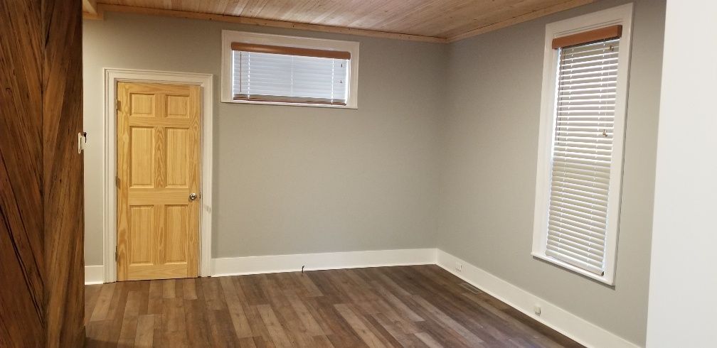 Empty room with wood floors, gray walls, light wooden door, and two windows with blinds.