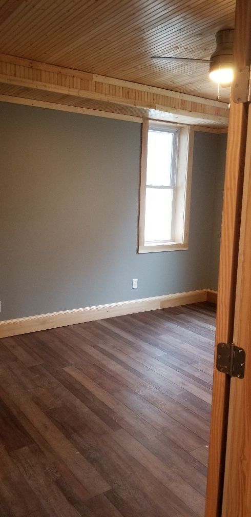 Empty room with gray walls, wood trim, and wood-look flooring. Light from window illuminates space.