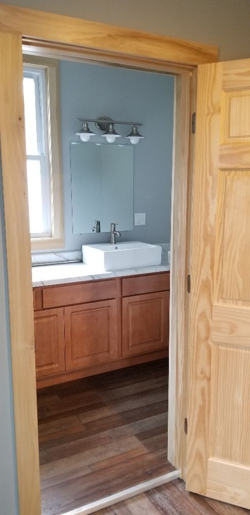 Open doorway into a bathroom with a sink, mirror, and light fixtures. Hardwood floors and wood cabinets.