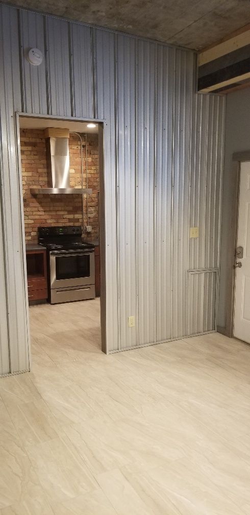 Interior of a room with corrugated metal walls, opening to a kitchen with brick.