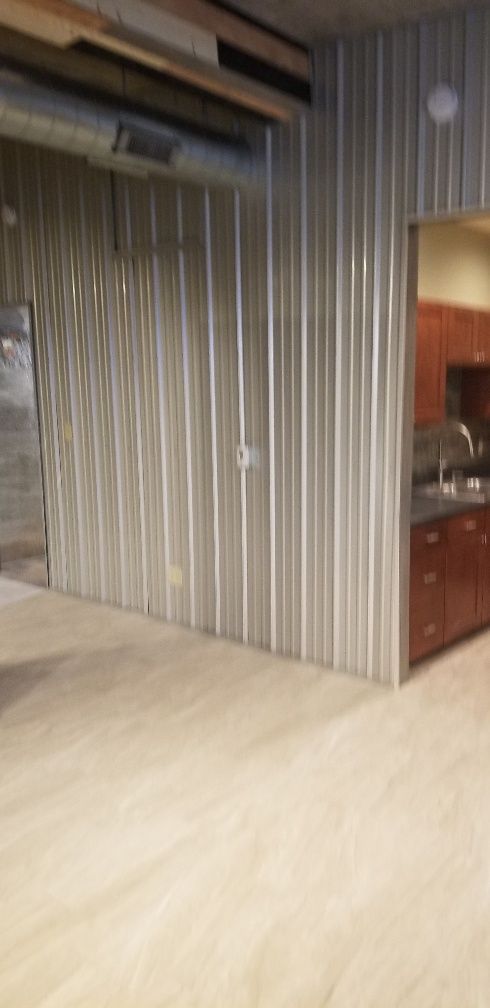 Interior of a room with corrugated metal walls and a kitchen area visible through a doorway. Light wood flooring.