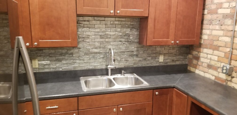 Kitchen with wooden cabinets, stainless steel sink, dark countertops, and stone backsplash.