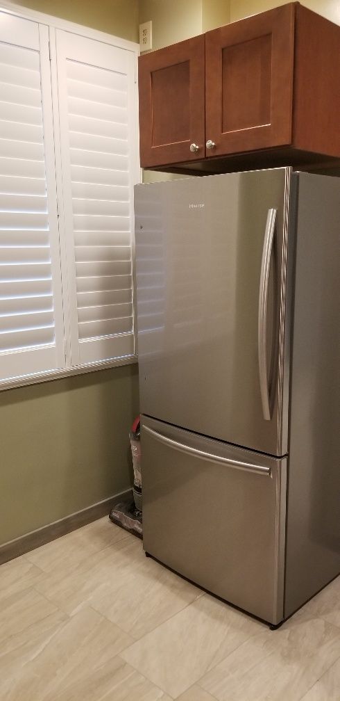 Stainless steel refrigerator in a kitchen with window shutters and brown cabinets.