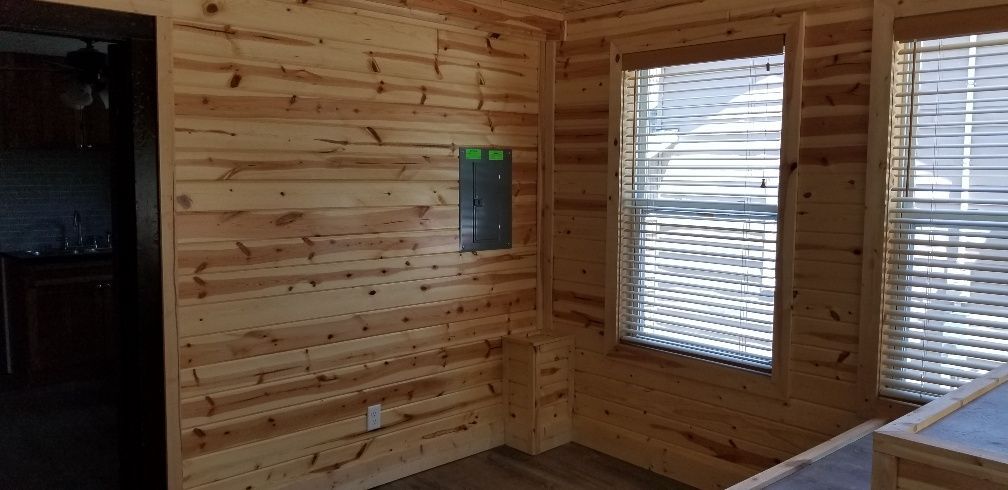 Interior of a room with wood paneled walls, a green electrical box, and windows with blinds.