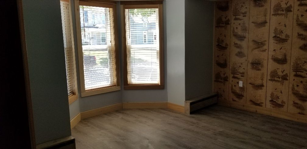 Room with light gray walls, wooden trim and flooring, and patterned wallpaper. Sunlight streams through windows.