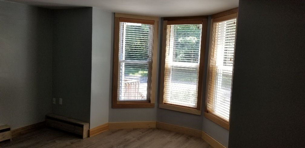 Room interior with a curved window, blinds, and wood trim. Pale blue walls and light wooden floor.