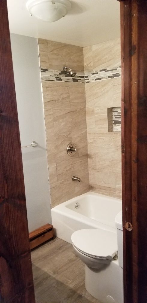 Bathroom with beige tiled shower over a white bathtub. Toilet to the right with wood trim on either side.