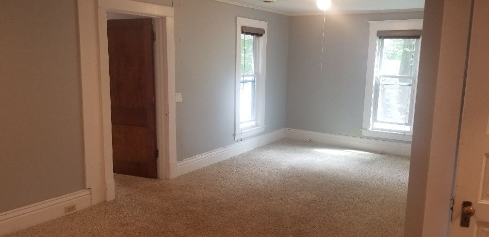 Empty room with grey walls, brown carpet, two windows, and a doorway with a brown door.