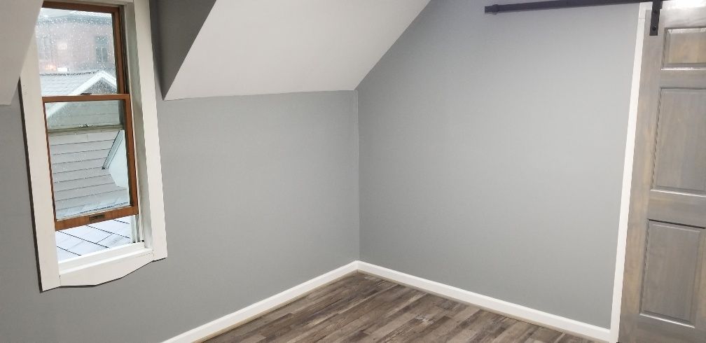 Empty room with gray walls, angled ceiling, wood flooring, and a window. A sliding door is on the right.