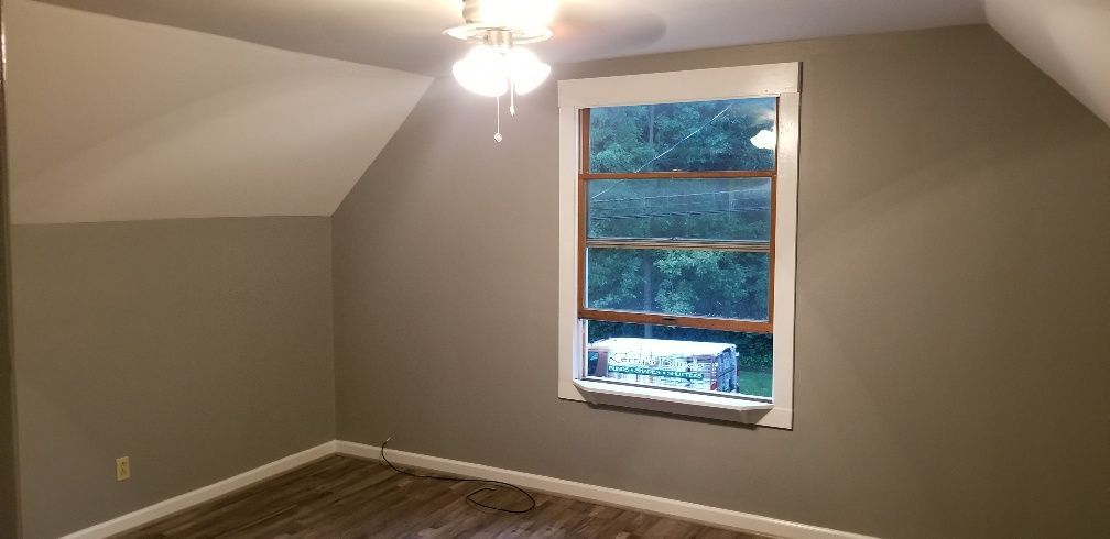 A room with a window, ceiling fan, and sloped ceiling. The walls are a dark tan color.