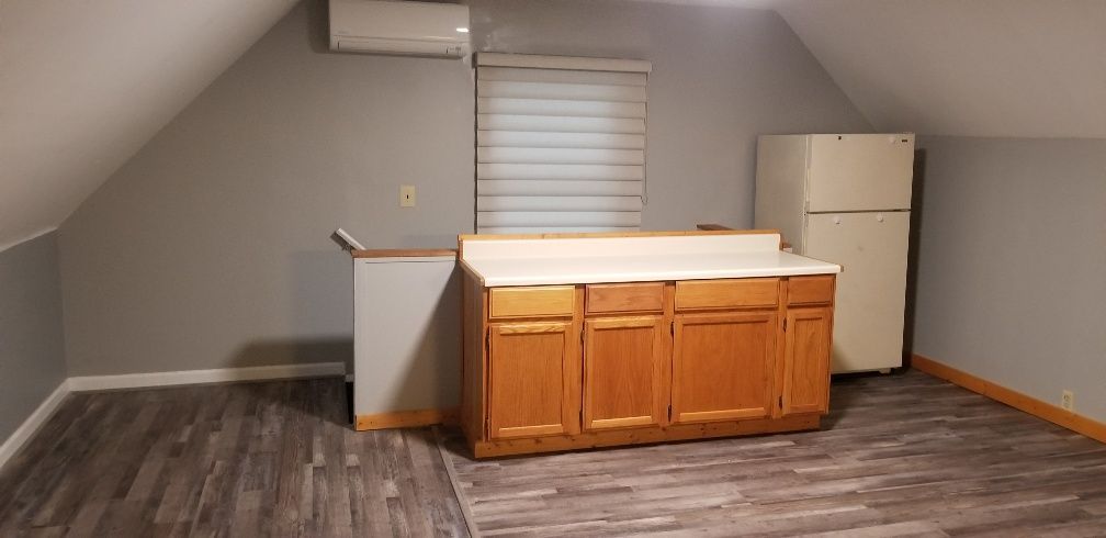 A small kitchen with wood cabinets, a fridge, and a window. The room has sloped ceilings.