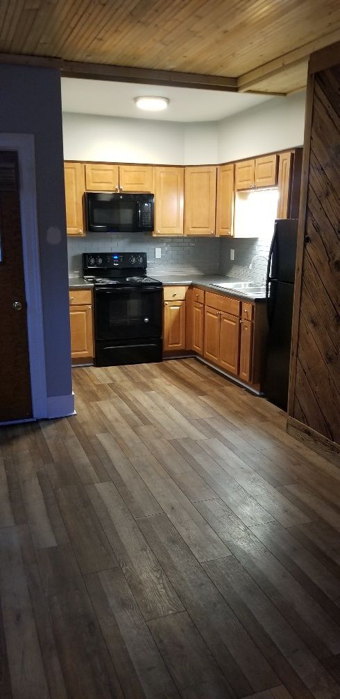 Kitchen with wooden cabinets and dark appliances, in a room with wood flooring and ceiling.