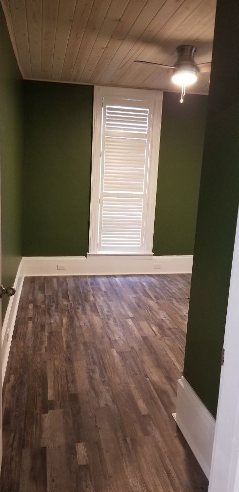A room with dark green walls, wood floor, white trim, window with shutters, and wood ceiling.