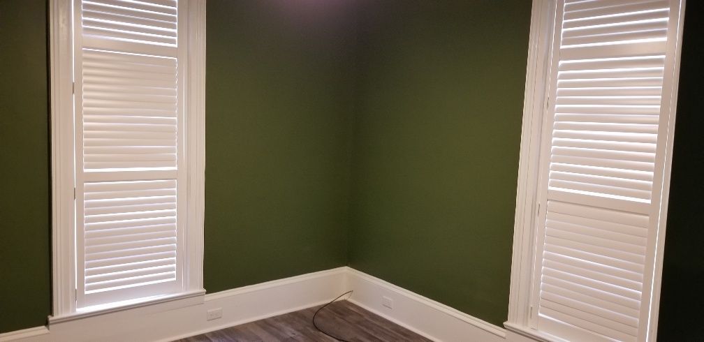 Corner of a room with green walls, two white shuttered windows, and a wood floor.