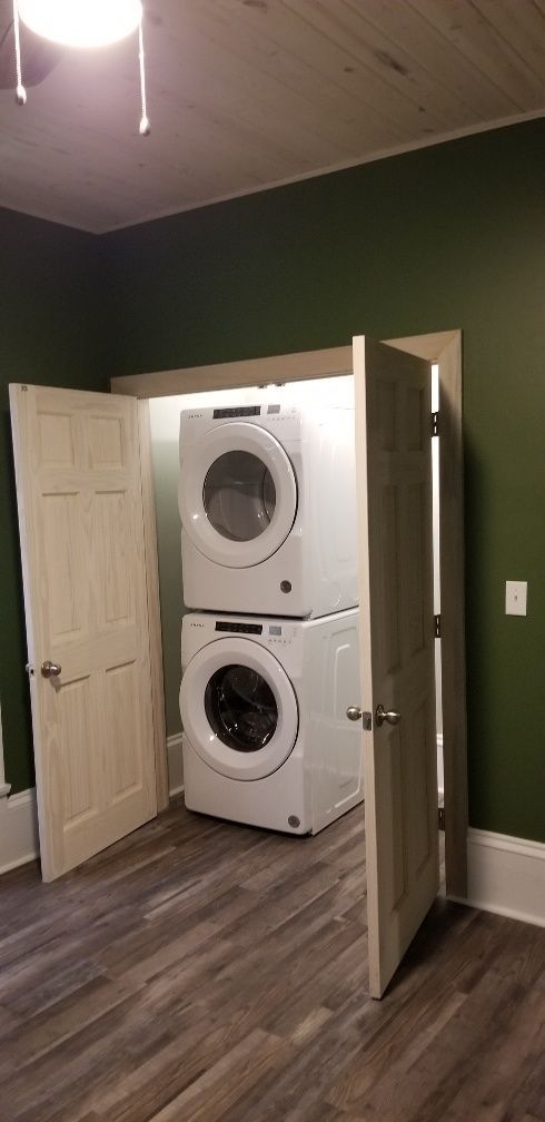 Stacked white washer and dryer in a closet, flanked by open doors, in a room with green walls and wood-look flooring.