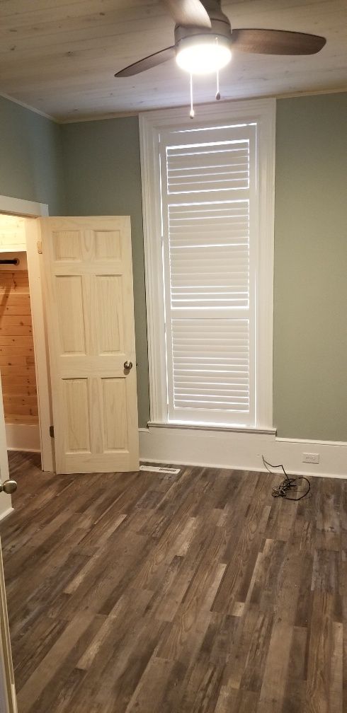 Bedroom with green walls, a tall window, and wood-look flooring. A ceiling fan hangs above.