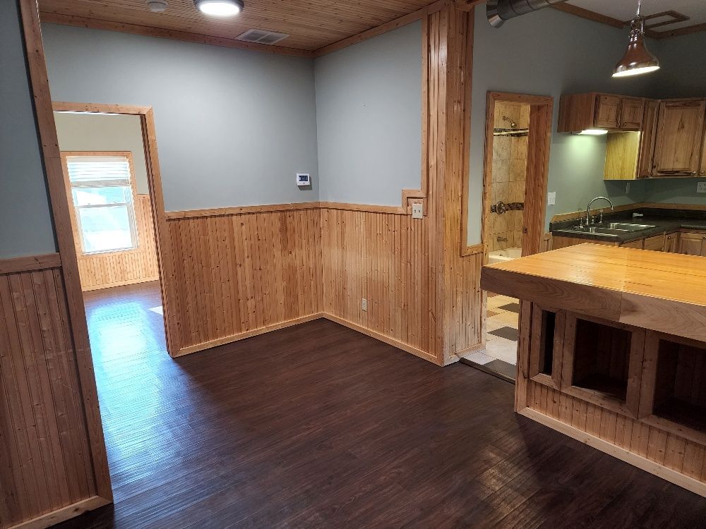 Interior room with wood paneling, dark flooring, kitchen area visible in background.
