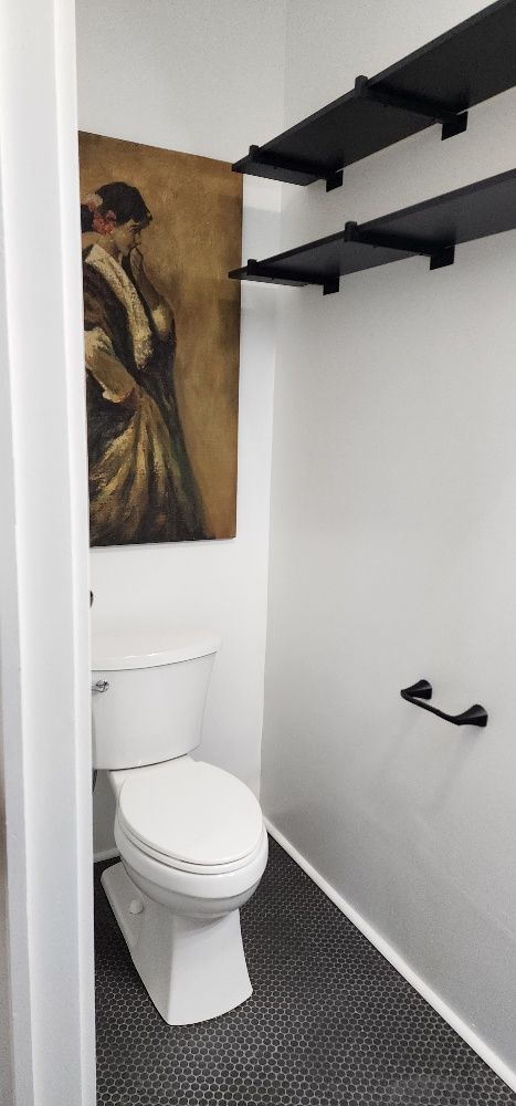 Small bathroom with black and white tile flooring, toilet, art, and black shelves.