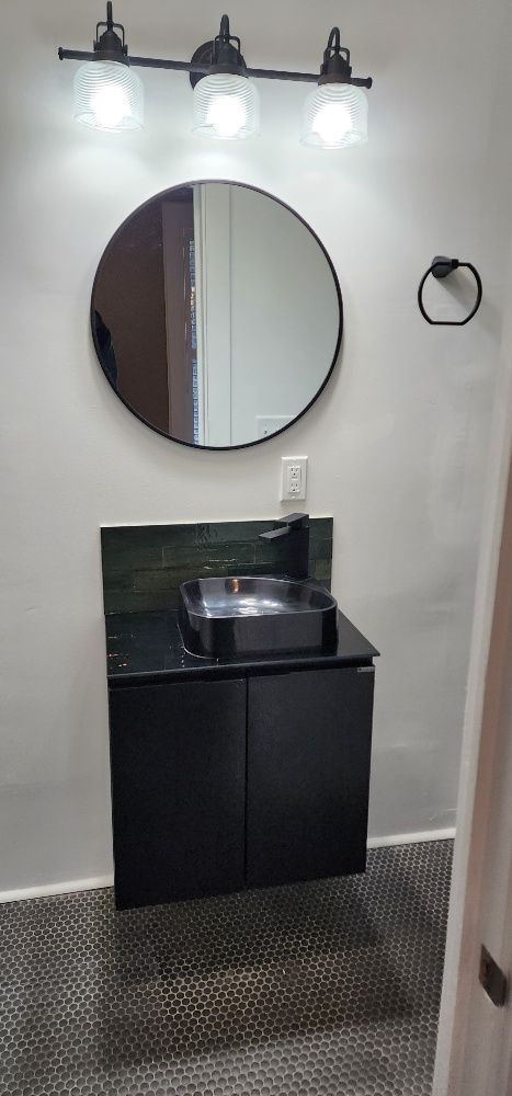 Black bathroom vanity with round mirror, lighting, and sink against a white wall.