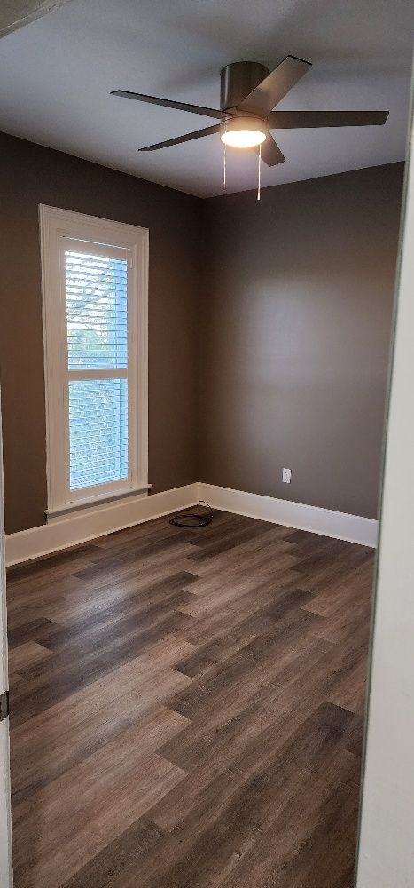 A bedroom with brown walls and wood-look flooring. A ceiling fan with a light is installed. Sunlight streams through a window.