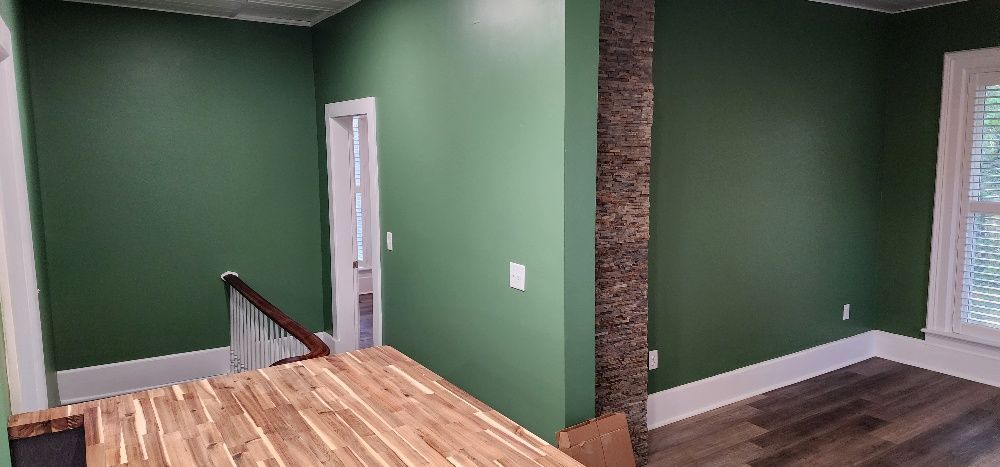 Green-painted room with wooden floors, a wooden table, and a textured pillar. White trim surrounds the walls and a door.