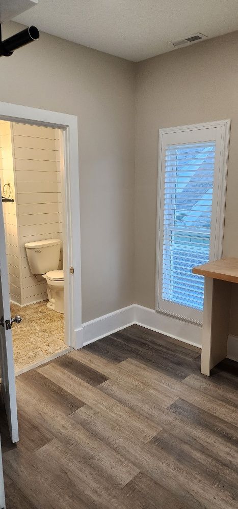 A room with wood-look flooring. A bathroom doorway is visible on the left. A window is on the right.