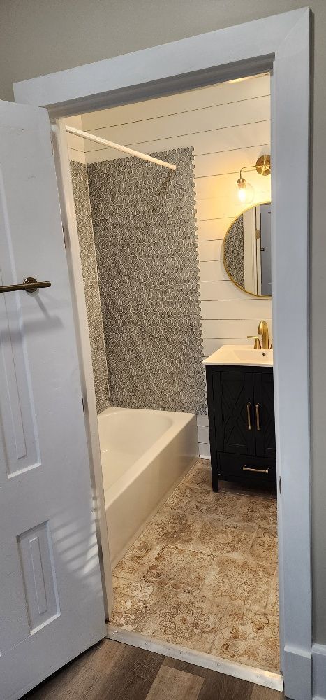 Bathroom viewed through an open white door. Features a pebble tile accent wall, black vanity, and gold fixtures.