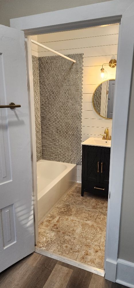 Bathroom interior seen through an open door, featuring a bathtub, vanity, mosaic wall, and round mirror.