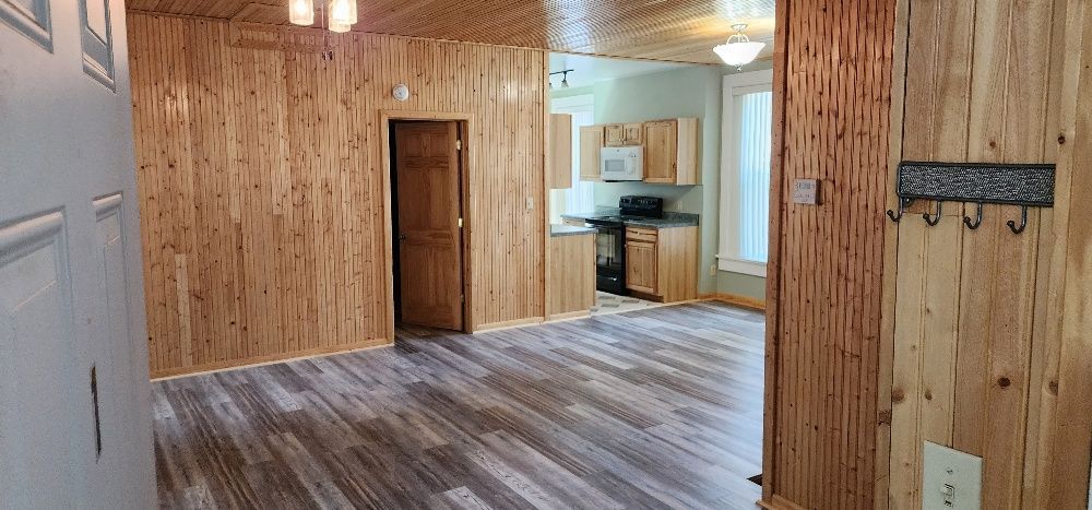 Interior of a room with wood paneling and gray flooring. Kitchen in the background.