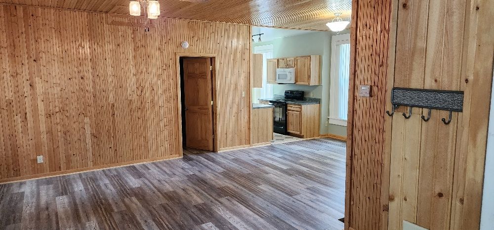 Interior of a room with wood paneling on walls and ceiling, kitchen in background.