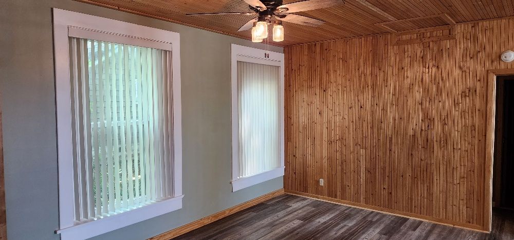 Interior room with wood paneling, two windows, and ceiling fan. Light-colored walls and flooring.