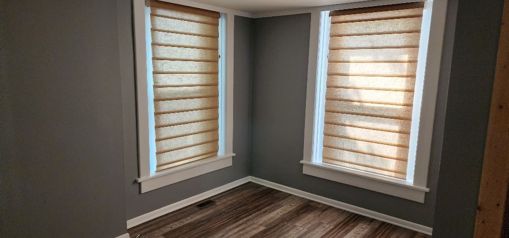 An empty room with two windows covered with light-colored blinds; gray walls and brown flooring.