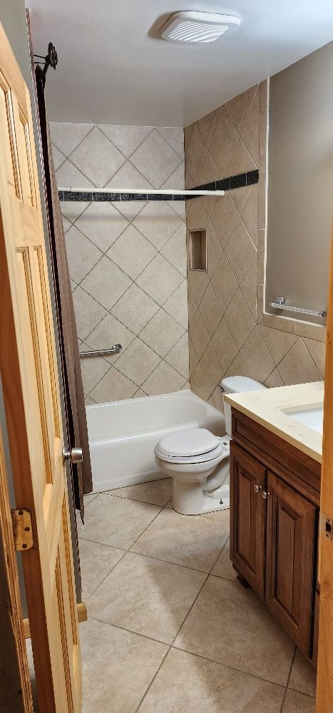 Bathroom with a tub, toilet, and vanity. Beige tiled walls and flooring. Wooden door is open in the foreground.